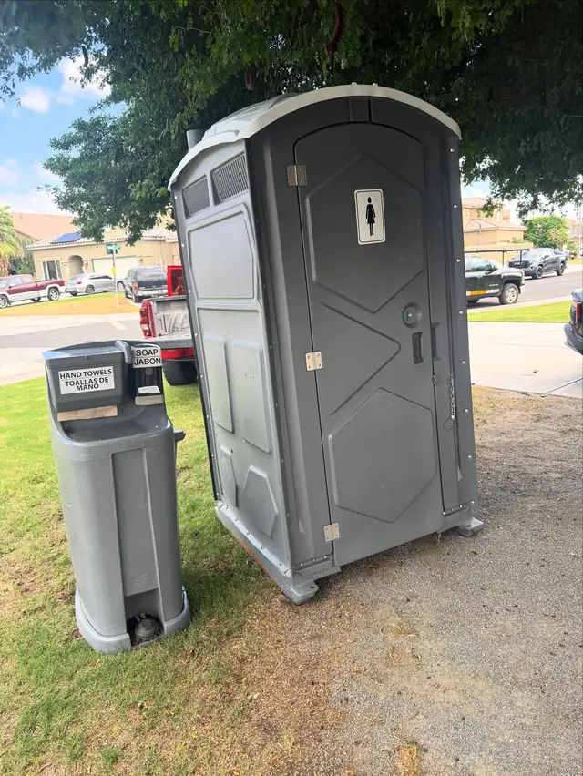 Gray porta potty on a lawn with a hand washing station beside it
